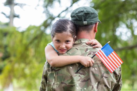A child smiling and hugging a service member in uniform who is holding a small American flag.